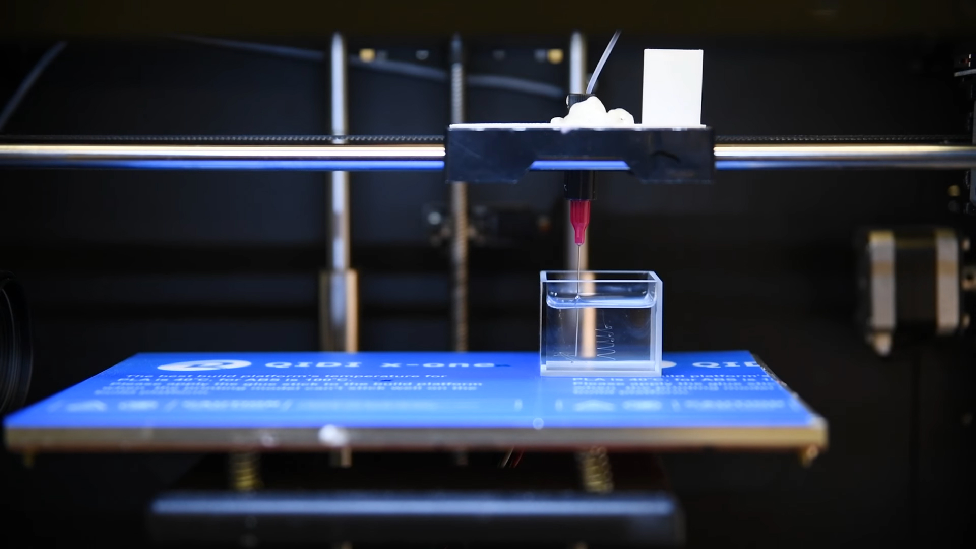 The modified Qidi X-One. A syringe and needle extrudes threads of water into a container of silicone oil. Photo via Berkeley Labs' YouTube.