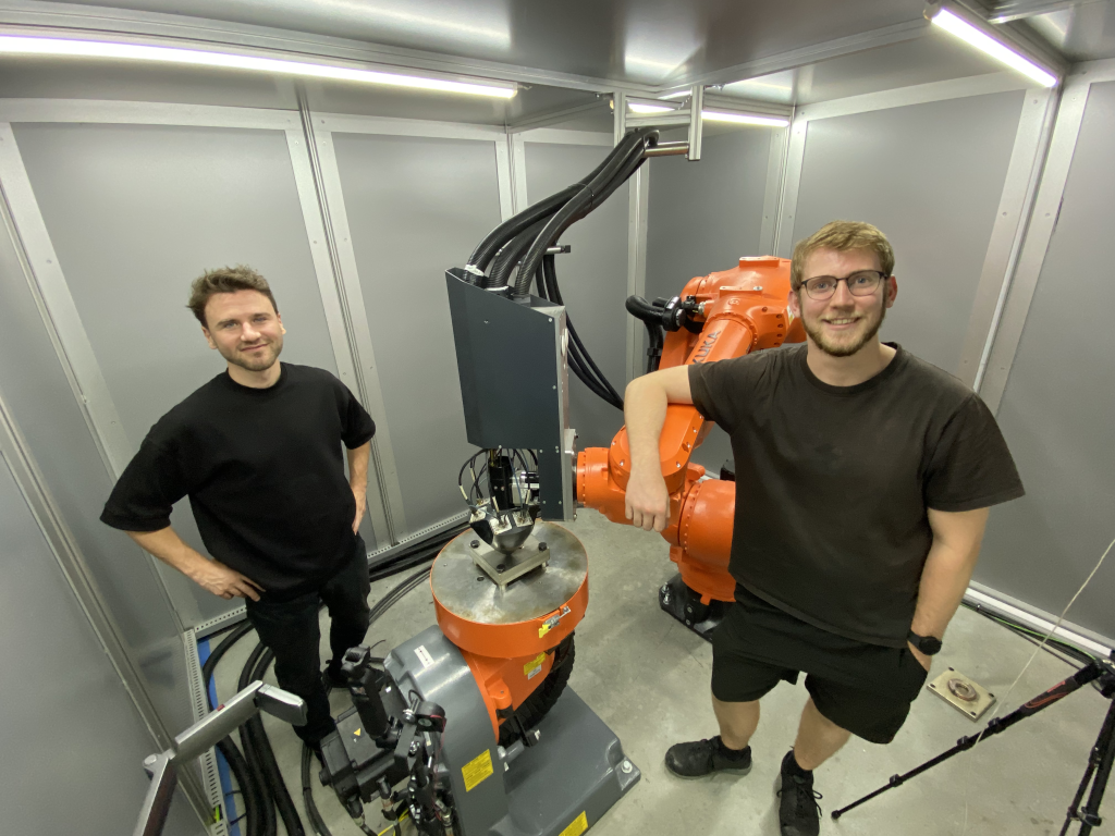 Daghan Cam (left) and Ai Build's Senior Design Enigneer Phil Barden with the company's Meltio DED 3D printer and Kuka robot. Photo by 3D Printing Industry.