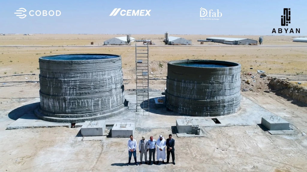 The world’s first on-site 3D printed large water tanks for chicken drinking water made for the Kuwait United Poultry Company. Representatives of Abyan, COBOD, and Kuwait United Poultry Company are seen in the front. Some chicken farms are seen at the top of the picture. Photo via Abyan.